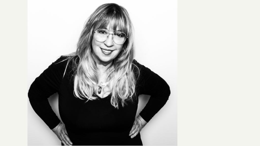A black and white portrait of a smiling woman with long hair and glasses, wearing a black top and standing confidently with her hands on her hips.